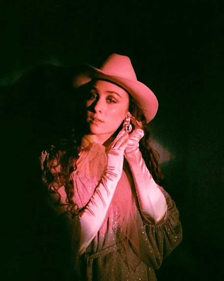 Una mujer con sombrero de vaquero rosa y guantes posa bajo una luz rosa.