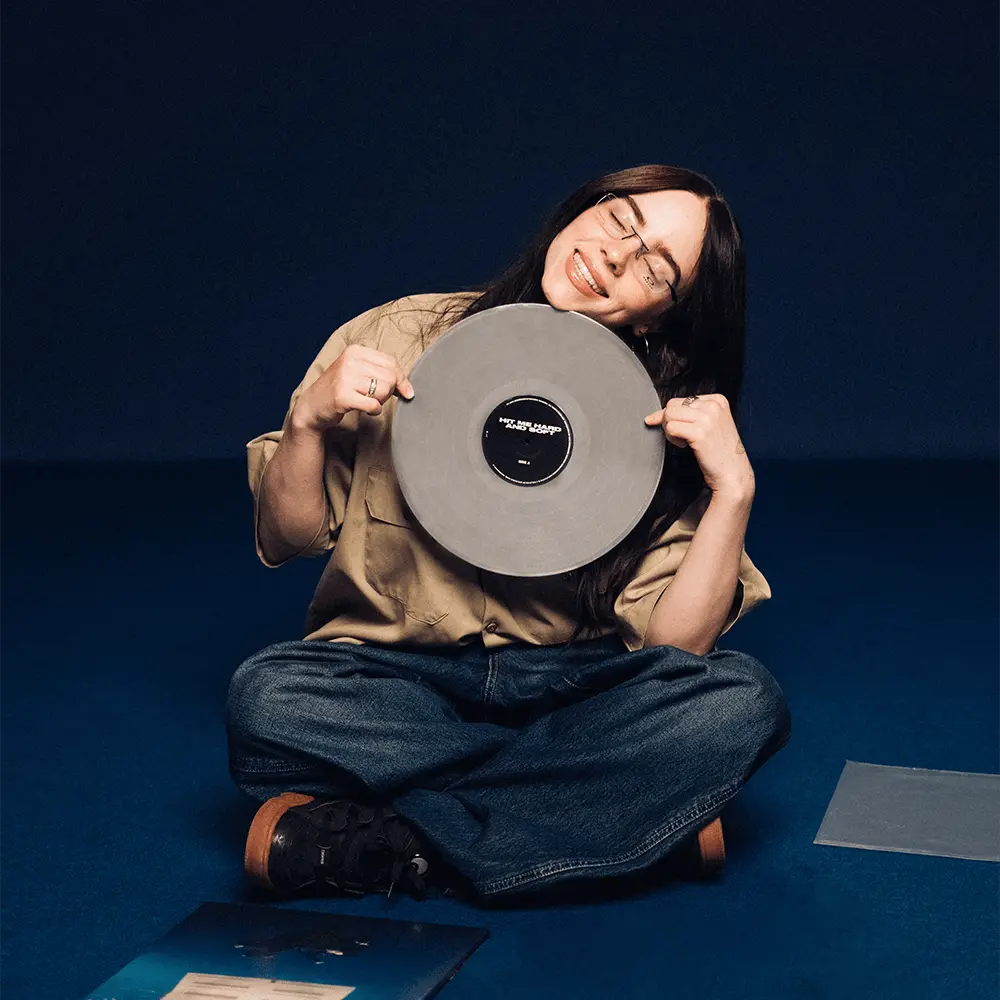 Billie Eilish sonríe mientras sostiene el vinilo gris de su álbum "Hit Me Hard & Soft".
