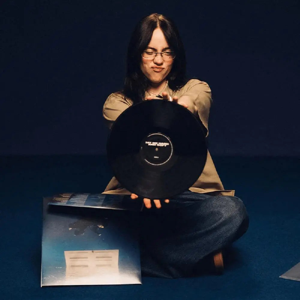 Billie Eilish holds up a black vinyl record with a gatefold album cover on the floor.